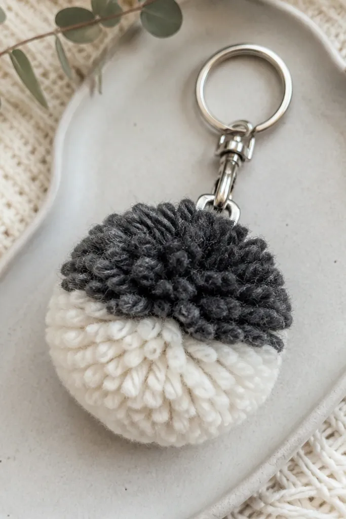 A pom-pom keychain made from soft sweater yarn in cream and charcoal. A small silver metal clip attaches it to a key ring.