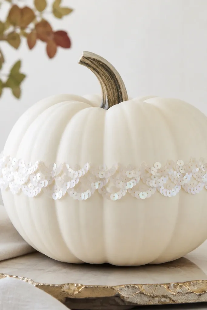 A white pumpkin with a scalloped trim band around the middle made from small white sequins. The sequins catch light in a gentle arc pattern.