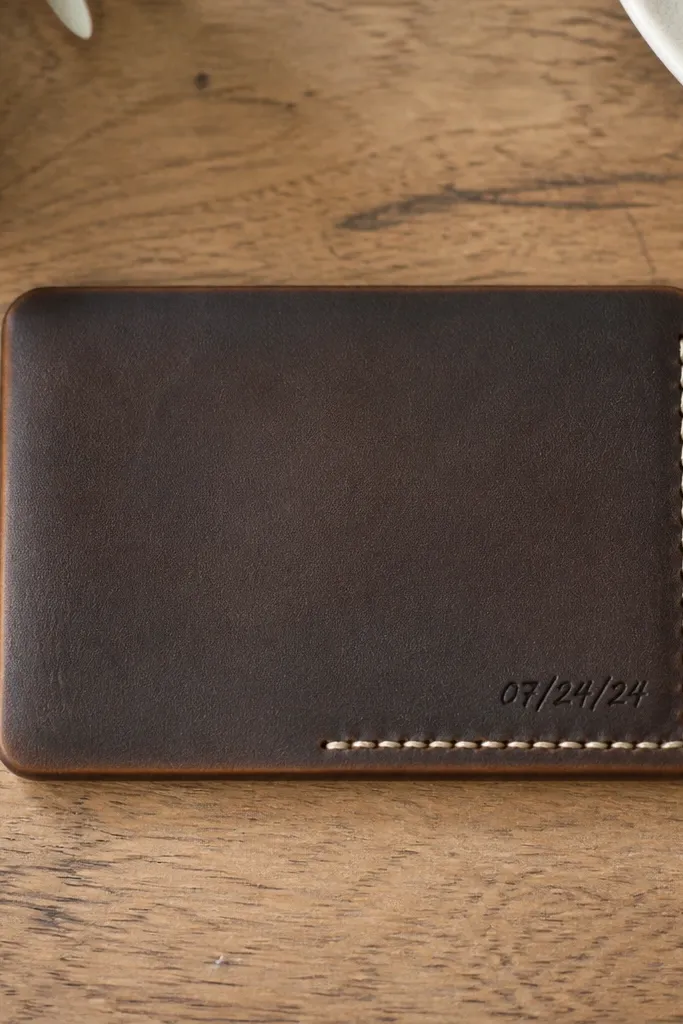 A slim leather wallet card shown flat on a desk. The leather is dark brown with a slightly burnished edge. In one corner there is neat stitching in a contrasting thread color, and a small stamped or handwritten-style date mark is visible. The card has rounded edges and a clean, minimal front.