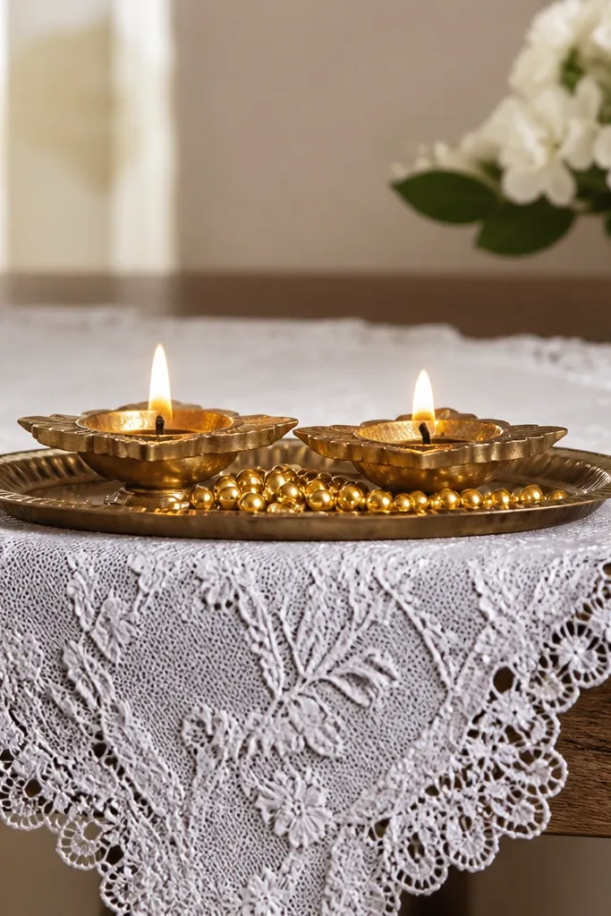 A white lace runner covers a table edge. Two diyas sit on a small tray, with gold beads scattered along the front edge of the tray.