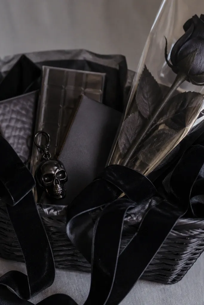A black basket with black tissue, a single faux black rose wrapped in cellophane, and velvet ribbon draped in layered loops. Inside are dark chocolate bars with minimalist black wrappers and a small skull keychain.
