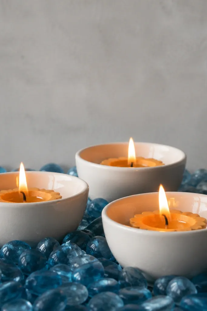 White ceramic candle cups each hold a diya. Around them is a bed of small blue glass pebbles. The background is a light grey wall and the flames look bright against the cool blue stones.