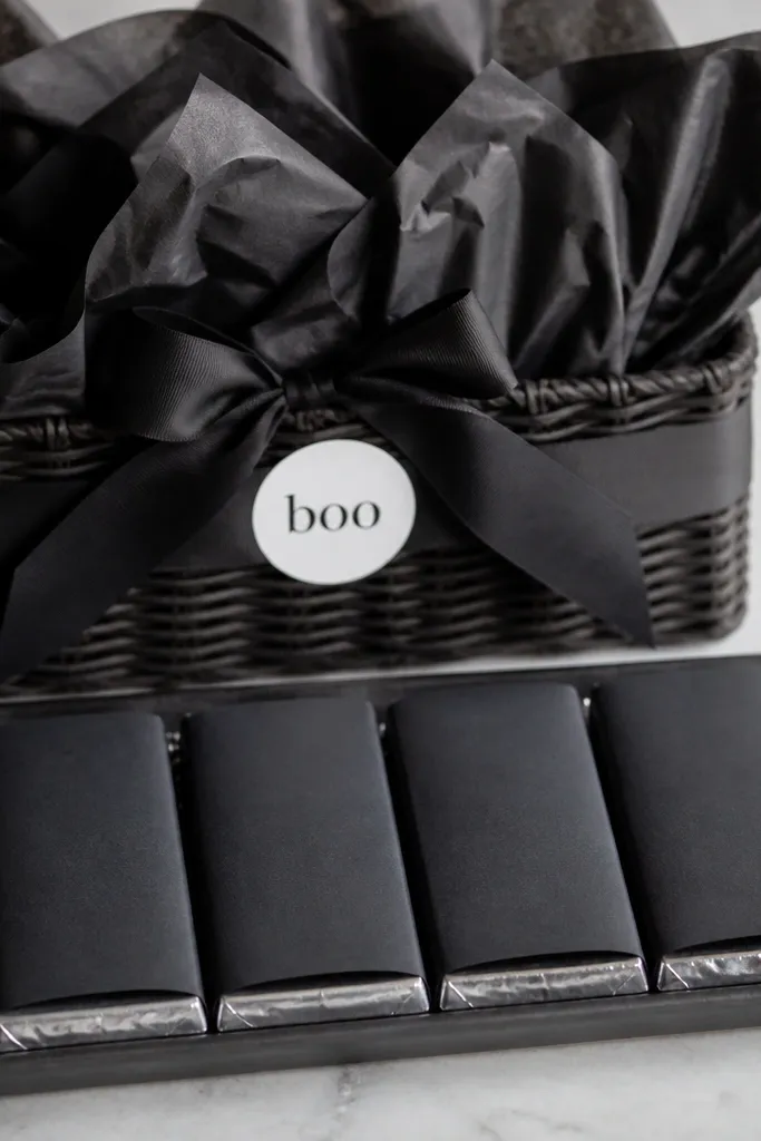 A basket with matte black tissue. A flat tray holds 4 black-wrapped chocolate bars arranged like a row. A small pinch of silver foil confetti sits around the tray edges, and a single white 'boo' label sticker is on the front ribbon.