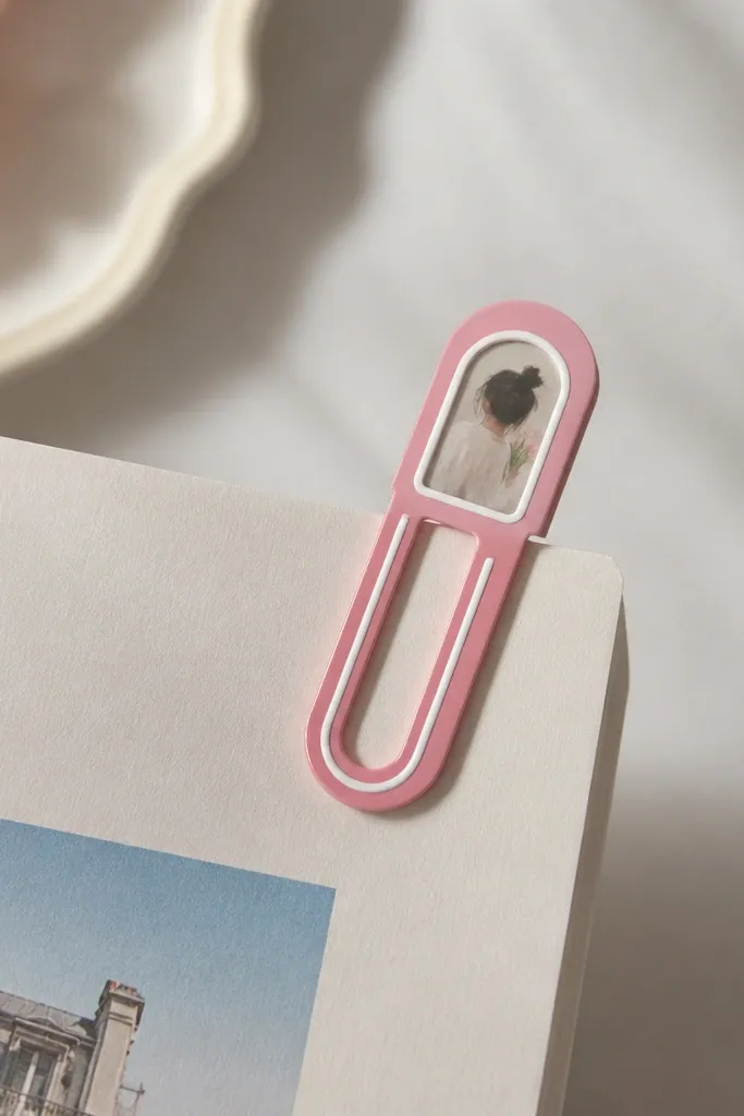 A metal paperclip-style bookmark painted in glossy pink with a thin white stripe. The clip has a small clear sticker window with a tiny photo inside near the top.
