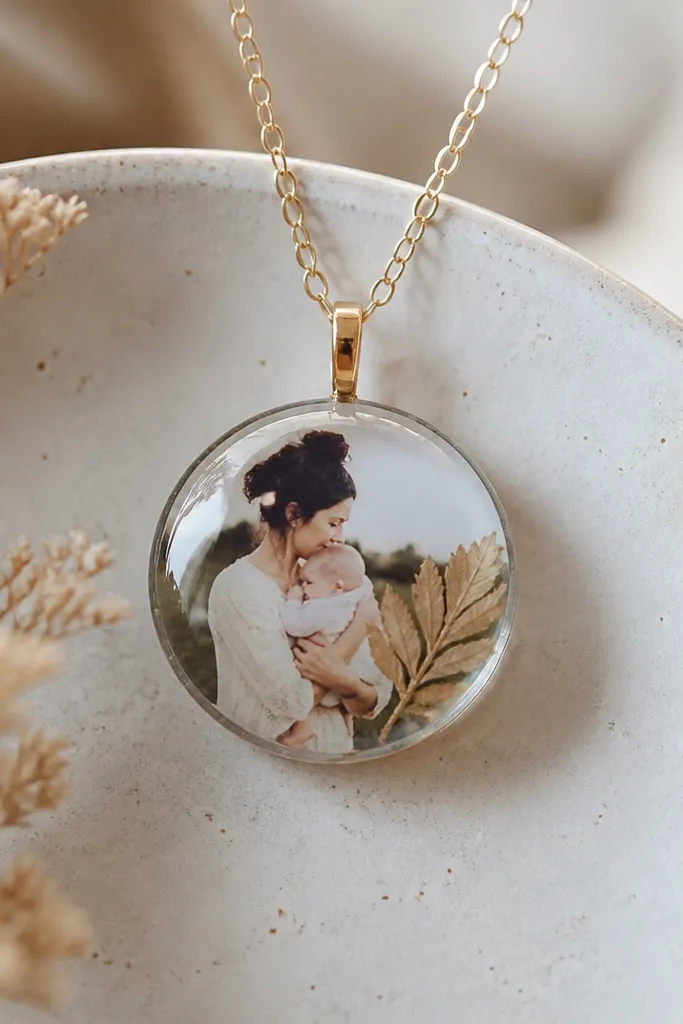 A small round pendant on a thin chain, with a tiny photo sealed in clear resin. A pressed dried leaf sits near the edge, and the pendant surface looks glassy.
