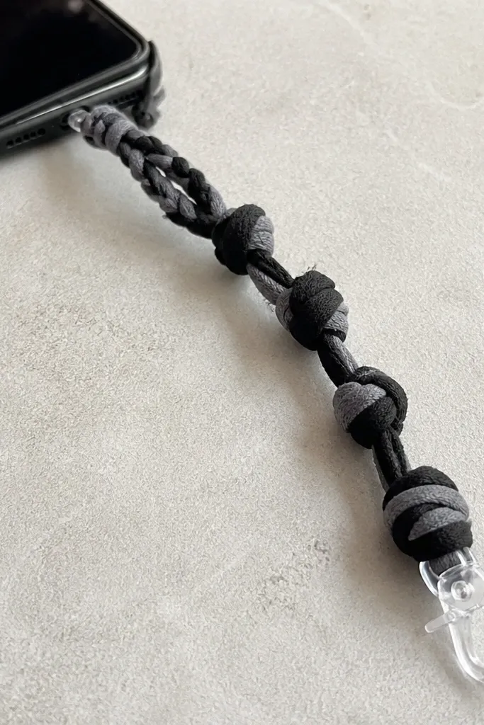 A close-up photo of a handmade phone strap made from strips of heather-gray and black T-shirt yarn. The strap has chunky knots every 1.5 inches, a braided section near the top, and a small clear plastic lobster-style clip at the end. The knots look raised and bumpy, with the yarn edges slightly fuzzy from cutting the old shirt.