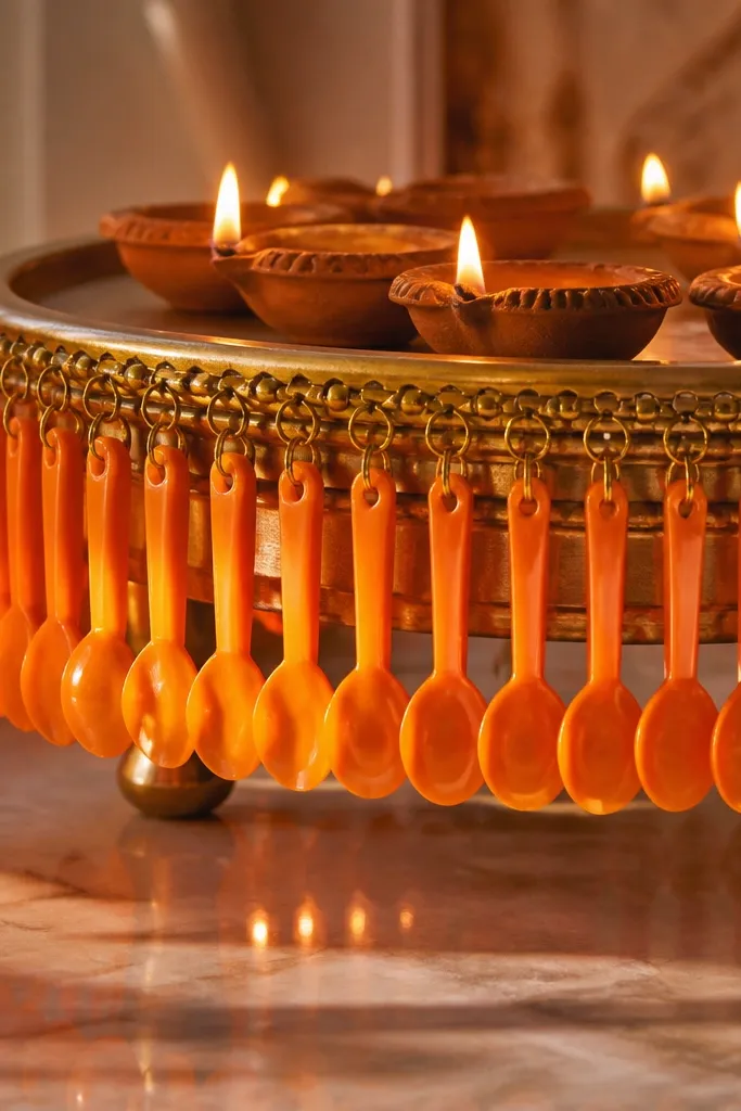 A fringe made from small plastic spoons painted orange hangs around a diya tray. Diyas sit beneath the fringe, and the spoons catch the flame light.