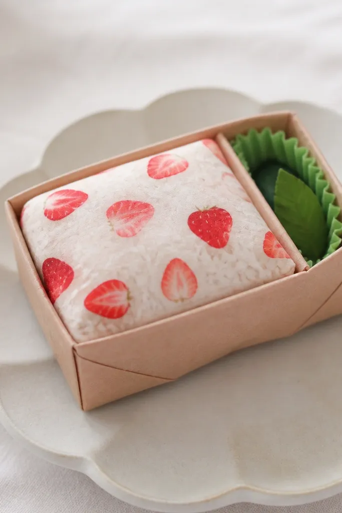 A small bento box-shaped paper squishy. The main top is pale rice with red strawberry pieces, and there's a side compartment with a tiny green garnish. The box has clean fold lines and a soft squishy top.
