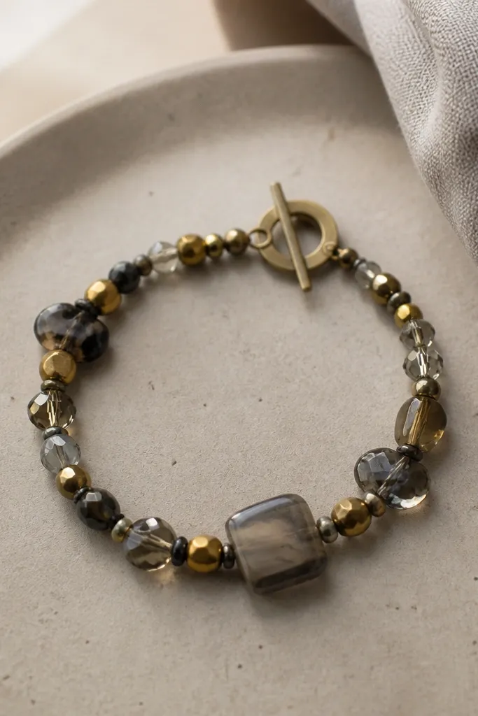 A bracelet made of mixed small beads and a few larger broken necklace pieces, strung on dark elastic with a simple toggle closure; the colors are muted gold and smoky gray.