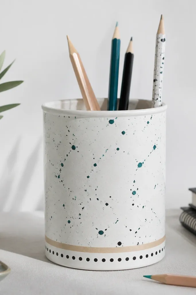 A cylindrical tin pencil holder painted matte white with scattered hand-made splatter dots in teal and black. Small polka dots line the lower edge, and a thin strip of masking tape defines the boundary.