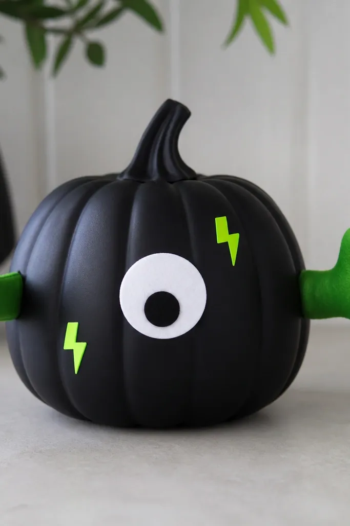 A black plastic pumpkin painted matte. Two small green felt hands reach from the sides like it's grabbing the viewer. A few neon sticker bolts and a simple white eye with a black pupil sit on the front.
