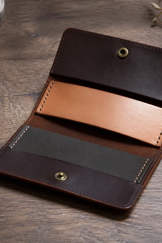 A small folded leather wallet-like strop case with a flap. Inside is a strip of smooth tan leather and a strip of darker leather on the other side, both stitched into place. The case has a simple brass snap and a visible burnished edge. The photo shows the strop leather surface catching light and the fold line staying crisp.