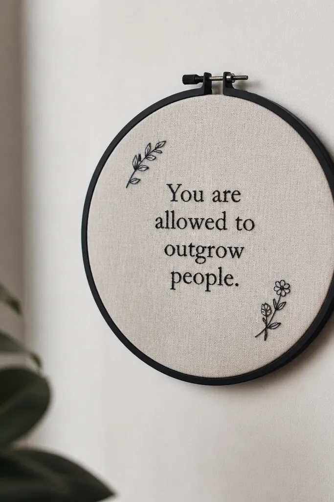 An embroidery hoop mounted on a wall with a small linen fabric inside. A quote is stitched in dark thread, with two tiny embroidered icons at the corners, and the hoop is painted matte black.