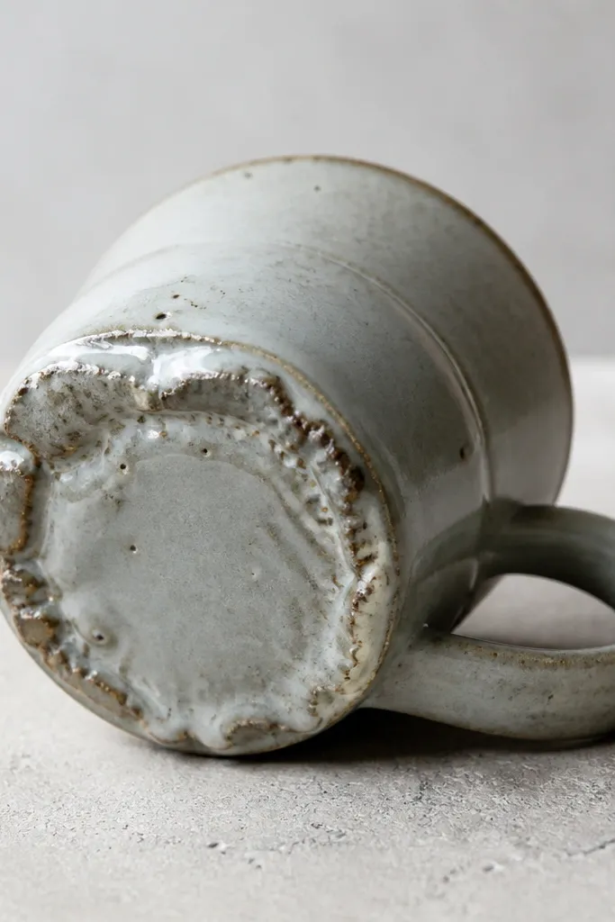 A mug photographed with a rough, broken-looking base ring where glaze pooled and fused to the kiln shelf. The bottom shows uneven glaze and a rough edge.