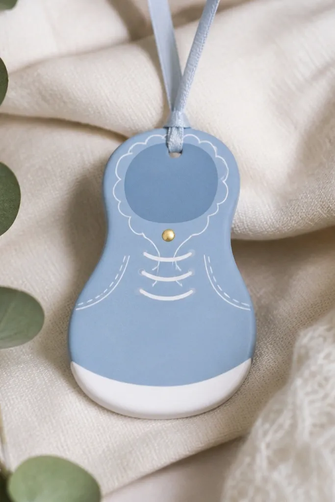 An oval ornament shaped like a baby bootie. The bootie is painted dusty blue with a white sole, tiny lace lines drawn with a fine paint pen, and a small gold dot near the top.