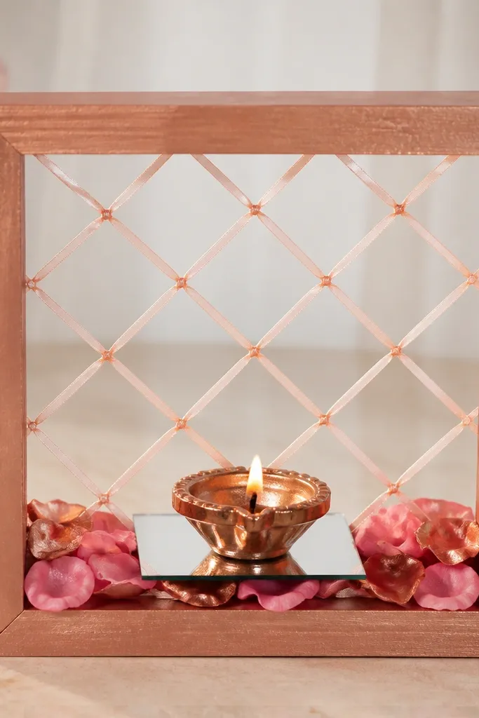 A small rectangular frame made from rose-gold painted cardboard stands on a table. Inside the frame is a lattice of thin ribbon, and a central diya sits on a mirrored tile. Pink and copper faux rose petals line the base.