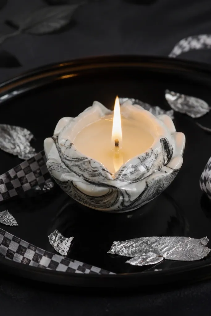 A glossy black lacquer tray holds a central white diya with marbled paint effect. Around it are thin strips of silver leaf and black-and-white check ribbon accents. The flame glows sharply against the dark background.