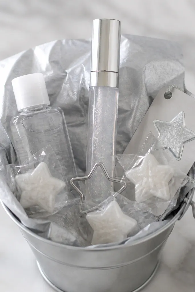 A small tin bucket with silver tissue paper. A tube of clear-with-sparkle lip gloss stands upright in the center, held by a mini star-shaped stand. Around it are star-shaped candies in clear wrap, a mini hand sanitizer, and a small silver "boo" sticker on a tag.