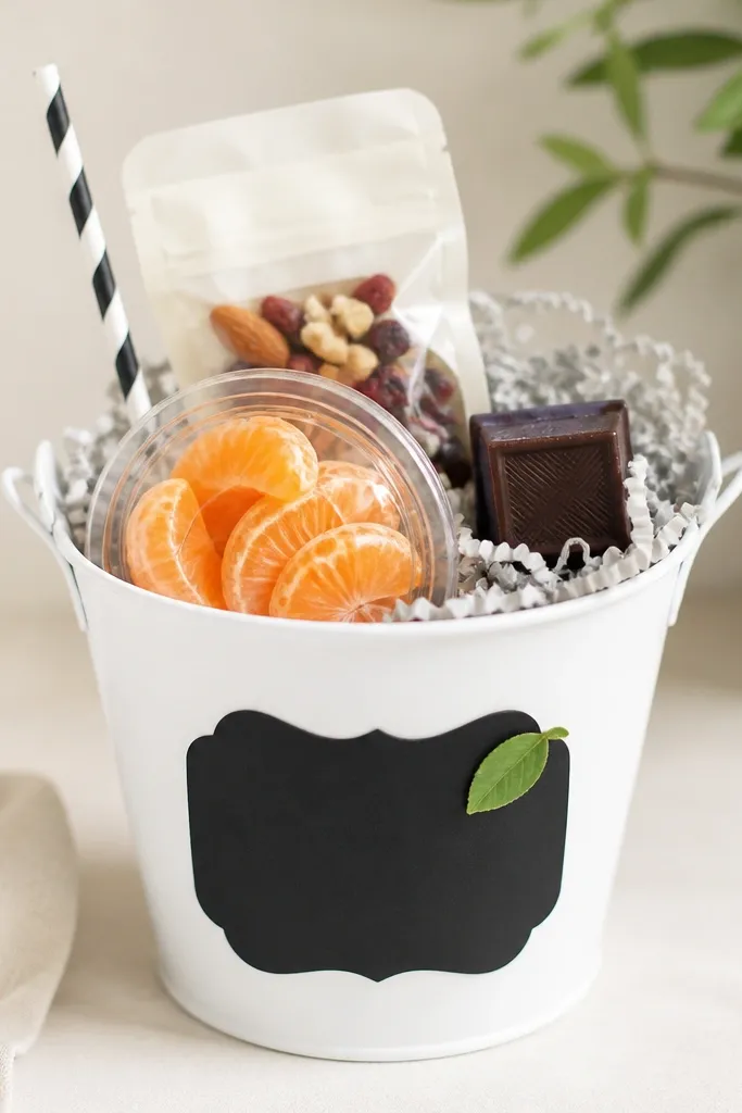 A white tin pail with a matte black chalkboard label on the front. Inside is pale gray shredded paper. There's a small sealed snack pack, a peeled mandarin in a clear snack container, and a mini dark chocolate square. The top has a black-and-white striped paper straw and one small green leaf sticker on the label.