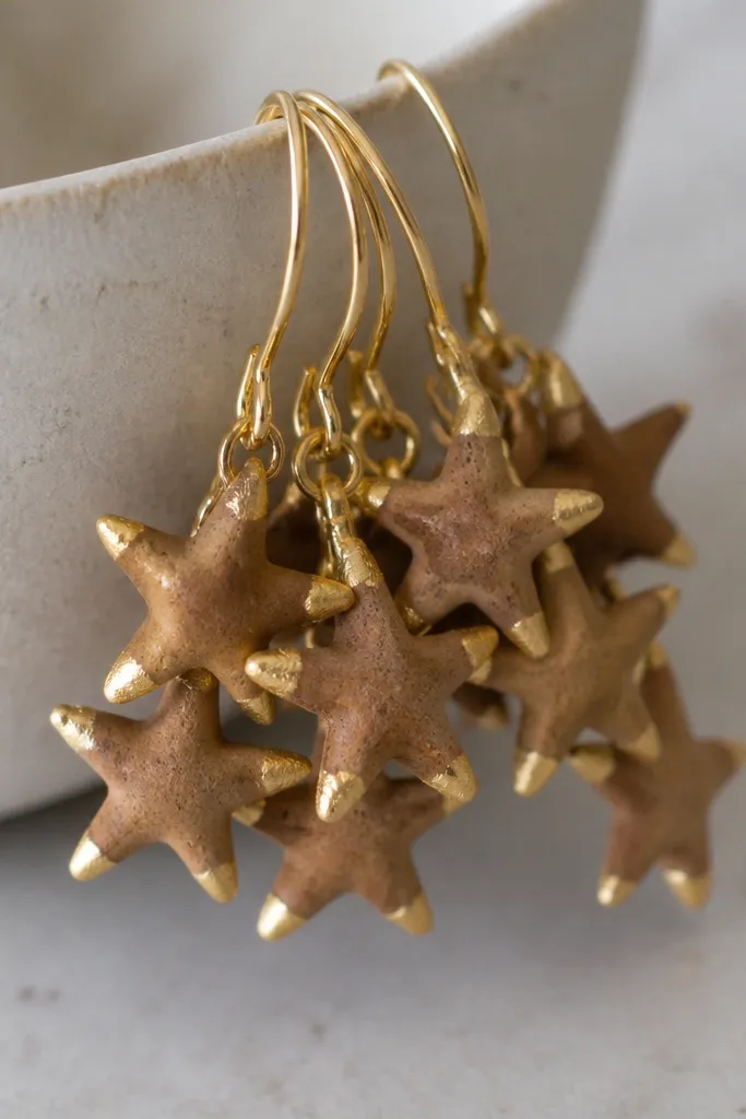 A cluster of tiny five-point stars. The stars keep their unpainted cinnamon-brown surface, and only the tips have matte gold paint or leaf. They hang from matching gold hooks.