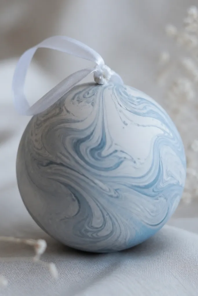 A round ornament with marbled swirls in light grey and pale blue, matte finish. The swirls look like ink pulled through paint. A white ribbon loop is tied at the top.