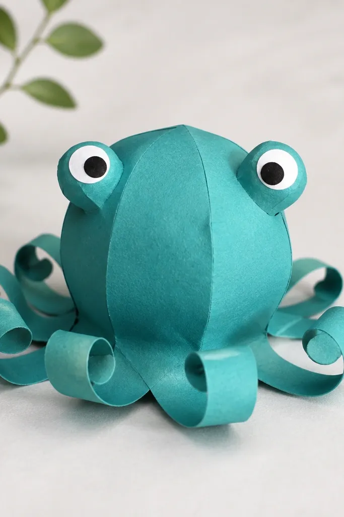 A teal paper squishy octopus with a rounded body and multiple curled tentacles at the bottom. The tentacles are thin strips curled into loops, each with a rounded end. The octopus has big eyes on stalk-like bumps made from folded paper.