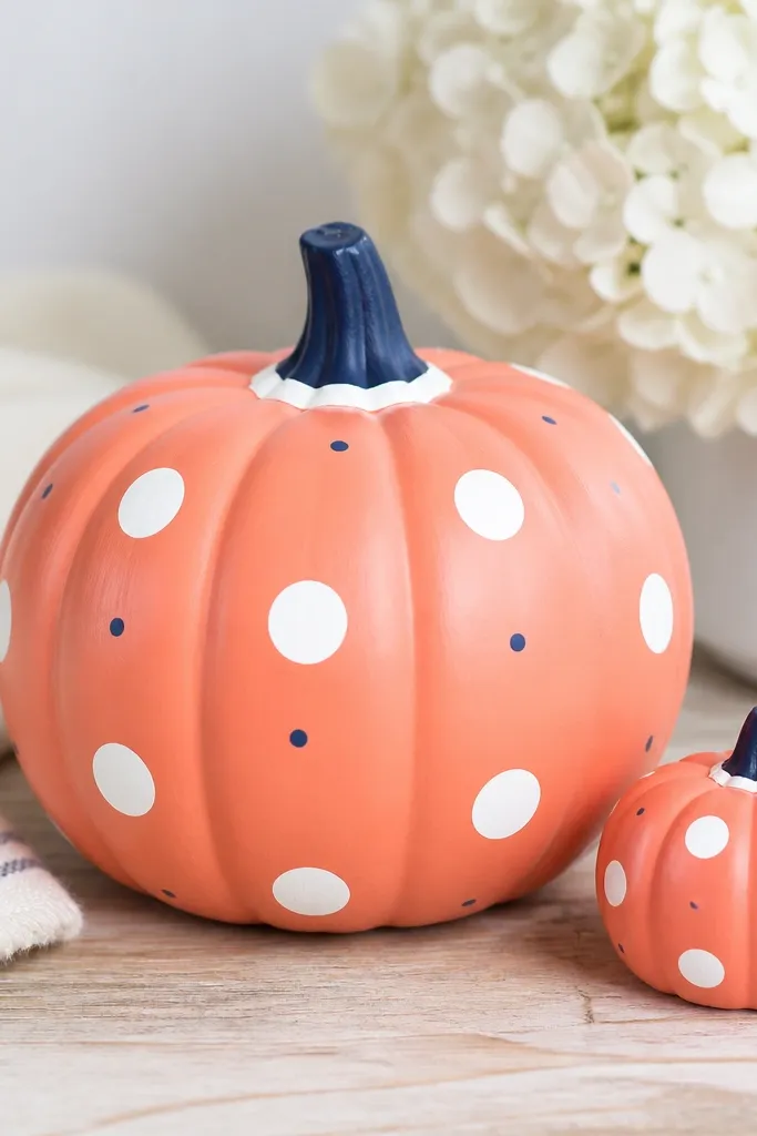 A bright coral pumpkin with white polka dots in two sizes, plus a few tiny navy dots scattered between; the stem has a navy cap and a thin white ring around the base.