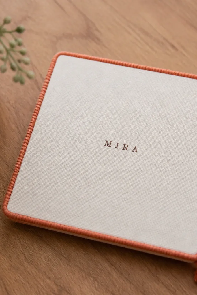 A small off-white canvas panel on a wooden table. The center has a stamped name in dark brown ink. A thin border of hand-wrapped embroidery thread runs around the edges in terracotta, with a tiny tassel at the bottom corner.