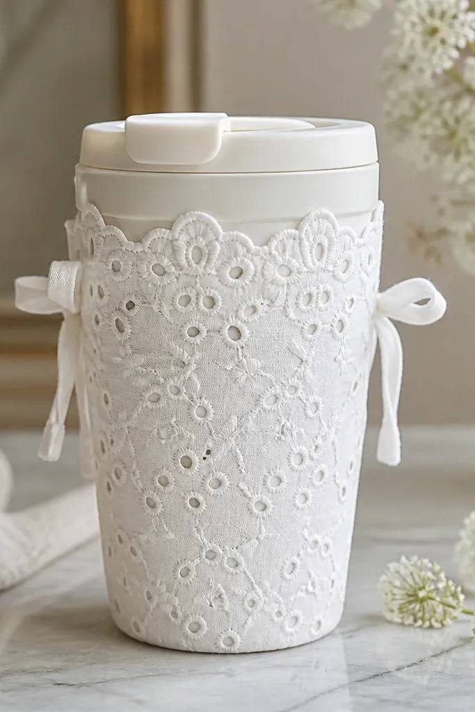 A travel mug covered in white eyelet lace fabric with a light cotton lining underneath. The sleeve has small ties at the sides and a scalloped top edge.
