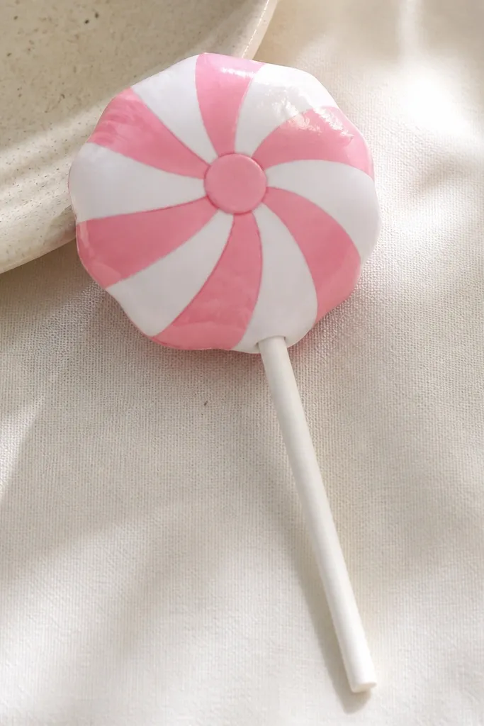A lollipop paper squishy with a round candy top wrapped in alternating pink and white strips. The stick is a thin paper strip, and the candy top looks slightly puffy. The wrap seams are hidden under a small top cap.