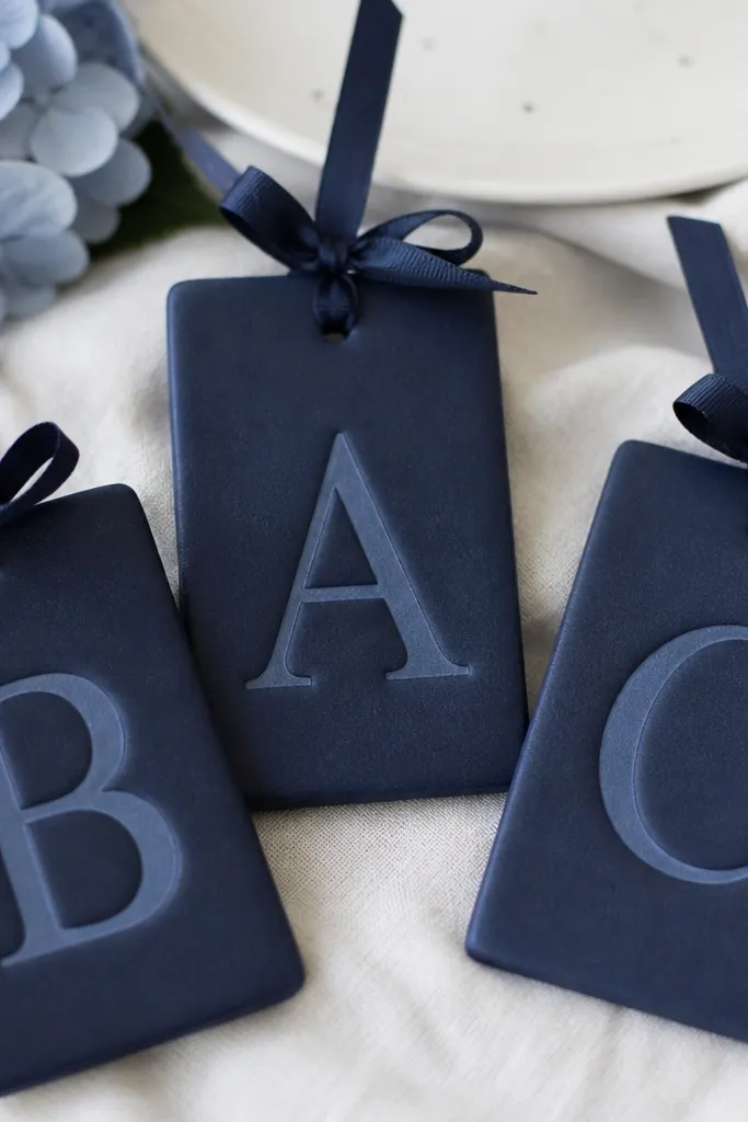 Rectangular salt dough tags with embossed or cut-out initials. The tag is deep navy, with a lighter navy monogram and a small bow tied at the top hole.