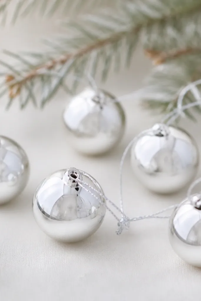 Round ornaments that look like small snowballs. They have a metallic silver finish with a smooth shine, and a subtle darker ring near the edge. A hole at the top holds a silver string.