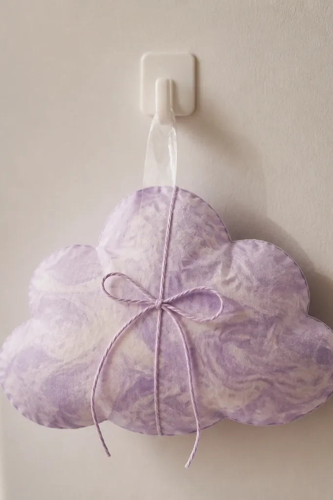 A puffy cloud-shaped paper squishy in lavender and pale purple with soft swirls, tied with a thin paper string bow. It hangs from a removable hook with a clear tape loop.