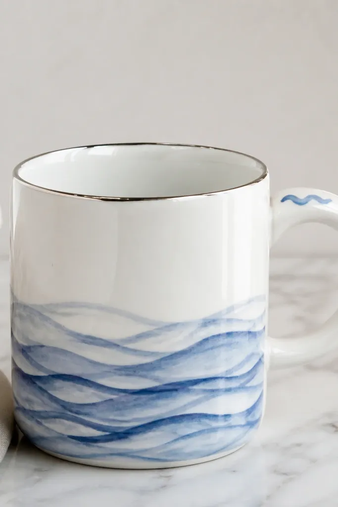 A white handmade mug with hand-painted blue wave lines across the lower half, like winter water. The rim has a thin silver metallic edge, and the handle is white with a small blue wave near the top. The glaze is glossy enough to show reflections of light.