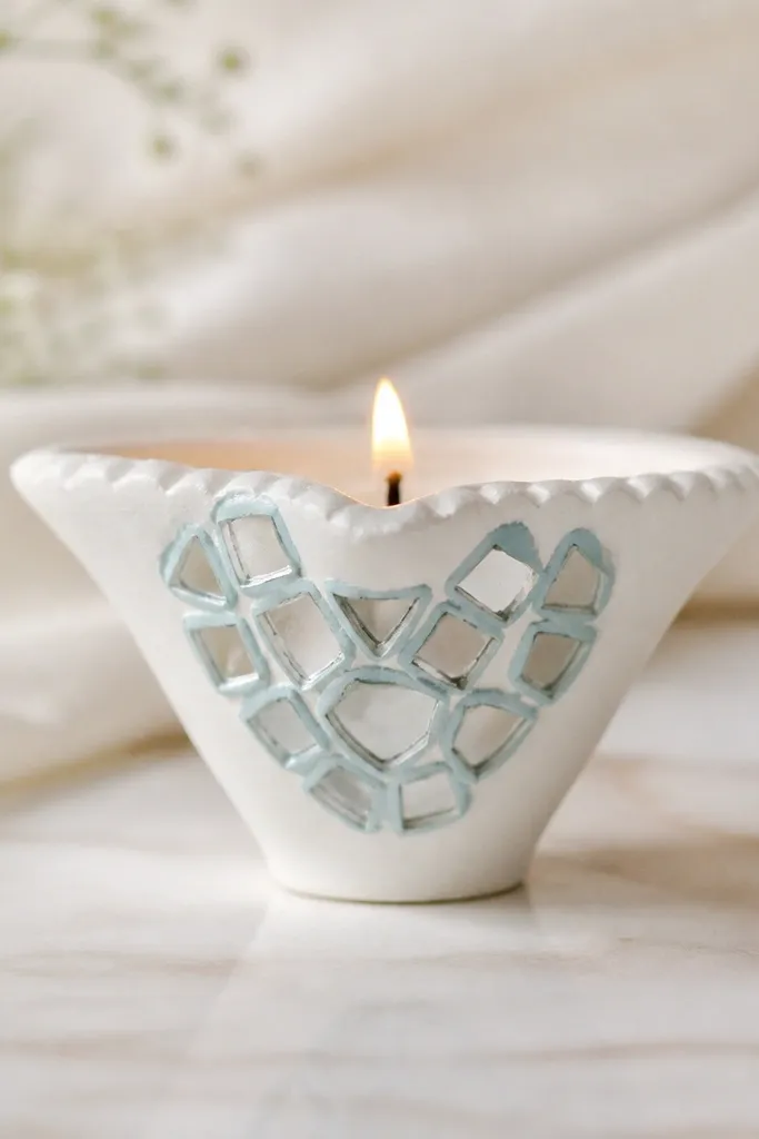 A diya painted white with small mirror pieces arranged in a heart-like cluster on the side. The mirror pieces are bordered with light blue paint so they look like sea glass.