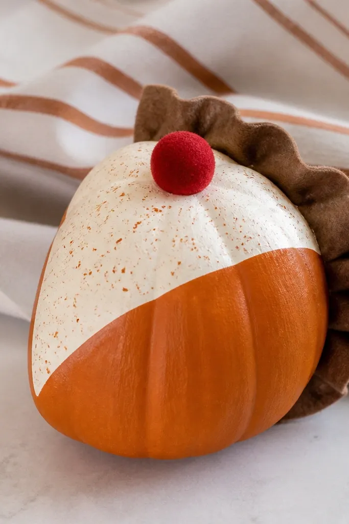 A pumpkin is painted to resemble a slice of pie. Brown felt forms a crust edge, and a small red felt cherry sits at the top. A cream filling area has tiny orange speckles, and the piece sits in front of a striped cloth.